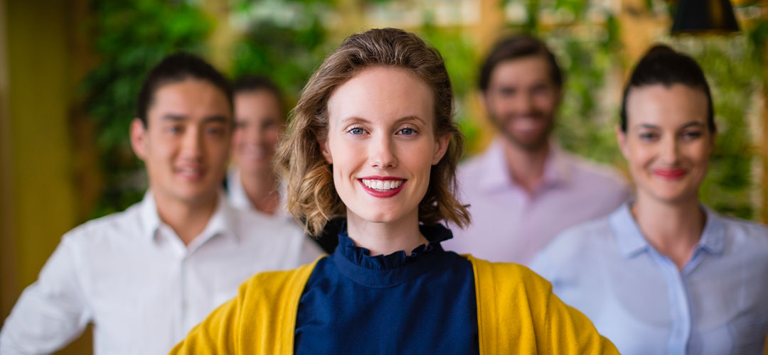 equipe-sourire-femme-happiness-manager Femme souriante au premier plan d'une équipe joyeuse, représentant la satisfaction des salariés bénéficiant d'un CSE externalisé avec des avantages adaptés